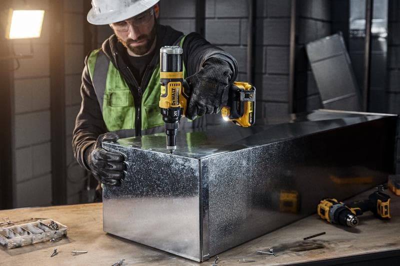 En person med skyddsutrustning använder ett DEWALT DCF403NT batteridrivet elverktyg för att arbeta med en metalllåda i en industriverkstad. Olika skruvar och verktyg syns på arbetsbänken.